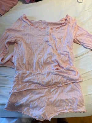 Ljusrosa pyjamas med volangdetalj - Söt ljusrosa pyjamas i två delar med långärmad topp och shorts. Toppen har volangkant vid halsen, tre knappar framtill och ett diskret ribbat mönster. Materialet ser mjukt och bekvämt ut, perfekt för mysiga kvällar hemma.