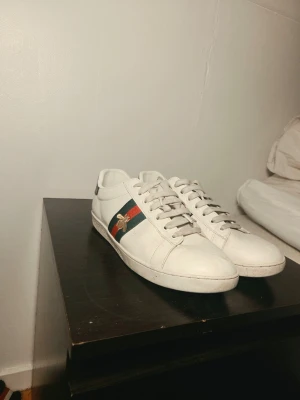 Gucci vita sneakers med bi-detalj - Säljer ett par vita sneakers från Gucci med klassiska gröna och röda ränder på sidorna samt en guldfärgad bi-detalj. Skorna har vita snören och detaljer i grönt och rött på hälen. Tillverkade i läder med gummisula. Perfekta för dig som gillar lyxiga och trendiga sneakers. Hör av dig innan du köper!