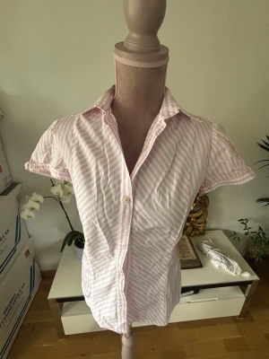Rosa och vit randig skjorta - Kortärmad skjorta med rosa och vita ränder. Skjortan har klassisk krage, knappar framtill och lätt figursydd passform. Perfekt för en avslappnad men ändå stilsäker look.