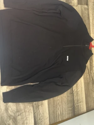 Svart tröja med dragkedja från BOSS - Svart långärmad tröja från BOSS med halv dragkedja framtill och diskret logga på bröstet. Tröjan har en enkel och stilren design, perfekt för en clean look. Tillverkad i mjukt material för extra komfort. Pris kan diskuteras 