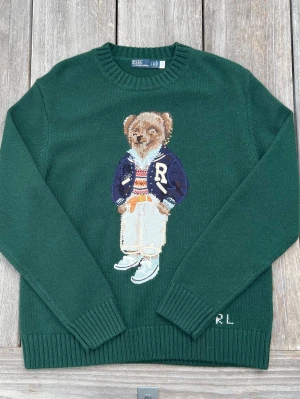 Ralph lauren bear stickad tröja  - Mörkgrön stickad tröja med rund hals och ribbade muddar. På bröstet finns ett stort motiv av en björn i preppy kläder och bokstäverna RL broderat vid nederkanten. Perfekt för dig som gillar unika och lekfulla plagg.