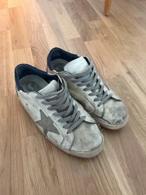 Vita sneakers med grå stjärna - Säljer ett par vita sneakers med grå stjärndetalj på sidan och grå skosnören. Skorna har mörkblå häl och är låga i modellen. Ovandelen verkar vara i läder och de har en klassisk, avslappnad look. Ett par väl använda Golden Gose, Box ingår tyvär inte. Och priset är inte hugget i sten så tveka inte att höra av dig vid intresse. Skorna är äkta.