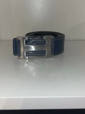Mörkblått skärp med H-spänne - Snyggt mörkblått bälte i skinn med en stor, silverfärgad H-formad metallspänne. Bältet har en stilren design och synliga sömmar längs kanterna för en exklusiv känsla. Perfekt accessoar för att lyfta vilken outfit som helst.