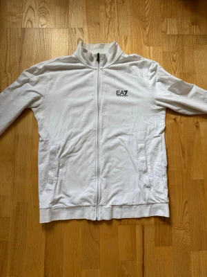 Vit zip hoodie från Armani - Snygg vit hoodie från Armani med dragkedja hela vägen och två fickor framtill. Tröjan har hög krage och EA7-logga på bröstet. Tillverkad i mjukt material som känns skönt mot huden. Perfekt för en clean och sportig look.