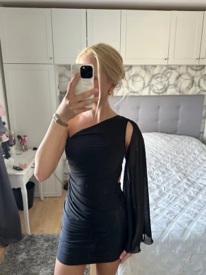 Svart oneshoulder kortklänning - Snygg svart kortklänning med oneshoulder-design och en lång, fladdrig ärm i transparent tyg. Klänningen har en tight passform. Storleken - S/M