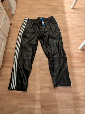 Svarta adidas byxor - Svarta adidas byxor med klassiska vita ränder längs sidorna. Byxorna har resår i midjan och är tillverkadi något slags fejkskinn material. Storlek XXXL. Men liten i storlek.