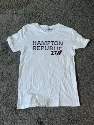 Vit t-shirt från Hampton Republic - Vit t-shirt från Hampton Republic med tryck 'HAMPTON REPUBLIC 27' på bröstet. Klassisk passform och rund halsringning. Perfekt till jeans eller shorts för en clean och enkel stil.