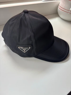 Svart keps från Prada i nylon - Snygg svart keps från Prada i glansigt nylonmaterial med klassisk böjd skärm. Kepsen har Pradas ikoniska metallogo på sidan och stilrena sömmar. Perfekt accessoar för en trendig och sportig look.