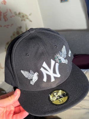 Svart New Era NY keps med broderi - Snygg svart keps från New Era med klassisk NY-logga framtill och broderade vita duvor på framsidan. Kepsen har rak skärm och sticker ut med sina detaljerade broderier. Perfekt för dig som vill ha en unik och trendig accessoar.