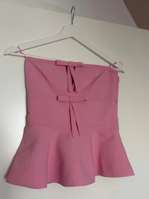 Rosa tubtopp med rosetter - Supersöt rosa tubtopp med tunna axelband och två dekorativa rosetter framtill. Toppen har en utsvängd nederdel som ger en feminin siluett. Perfekt för dig som vill ha en gullig och trendig look.