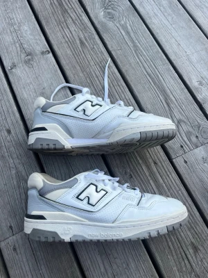 Vita New Balance 550 sneakers - Snygga vita New Balance 550 sneakers med svarta och grå detaljer. Klassisk låg modell med perforerade partier och robust sula. Skorna har tydlig New Balance-logga på sidan och är tillverkade i en mix av läder och syntetmaterial. I storlek 41,5