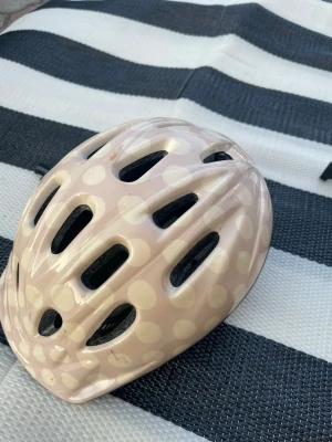 Cykelhjälm med prickigt mönster - Säljer en ljusrosa cykelhjälm med vita prickar och flera ventilationshål. Hjälmen har justerbart spänne och är tillverkad i plast med skuminsida för extra skydd. Perfekt för cykelturer och annan utomhusaktivitet.