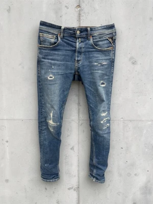 Replay jeans anbass - Säljer ett par blå  jeans med slitningar och hål på både fram- och baksida. Jeansen har klassisk femficksdesign, låg midja och är tillverkade i denim med stretch. Perfekta för en avslappnad och trendig look. Lite halv trasigt vid höger bak men inget som sticker ut så, hör av dig vid frågor Ai har beskrivit varan så var gärna nogrann med att höra av dig om du har minsta fundering 🌟