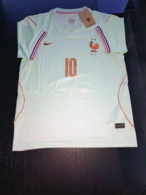 Vit Frankrike fotbollströja nummer 10 - Snygg vit fotbollströja med nummer 10 och Frankrikes landslagsemblem på bröstet. Tröjan har orange detaljer, Nike-logga och ränder i blått, vitt och rött vid axlarna. Perfekt för fotbollsträning eller match.
