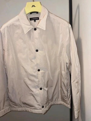 Beige jacka  från J.Lindeberg - Stilren beige jacka från J.Lindeberg med klassisk krage och svarta knappar framtill. Jackan har en minimalistisk design och är tillverkad i ett lätt, syntetiskt material. Perfekt för dig som gillar enkel och clean stil.