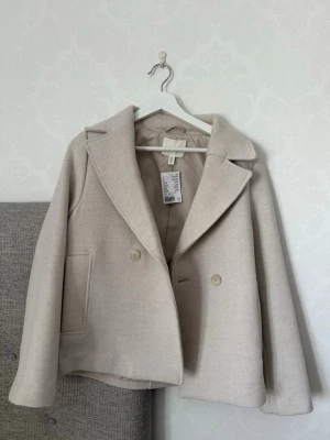 Beige kort kappa med bred krage - Stilren beige kappa med bred krage och dubbelknäppning framtill. Modellen är kort och har raka ärmar samt två stora fickor fram. Kappan är fodrad och har en mjuk, lätt strukturerad yta. Perfekt för dig som gillar minimalistisk och trendig stil.