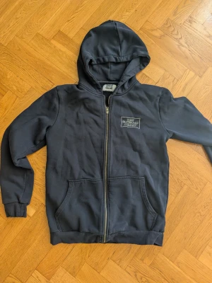 Mörkblå hoodie Sweet Skateboard - Mörkblå hoodie med dragkedja från Sweet Skateboard Company. Klassisk modell med huva, två fickor framtill och vit logga på bröstet. Skön och avslappnad stil, perfekt för skate eller chill. Tillverkad i mjukt bomullsmaterial.