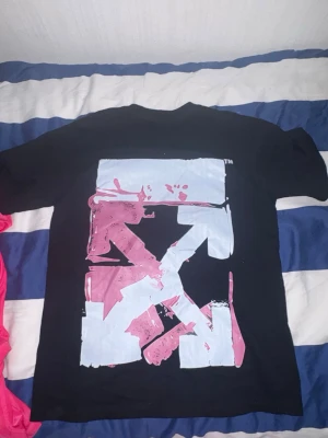 Off-White t-shirt - Svart t-shirt med stort grafiskt tryck i ljusblått och rosa på ryggen samt mindre Off-White-logga och handmotiv på bröstet. Klassisk passform och rund hals. Perfekt för dig som gillar streetwear och unika prints.
