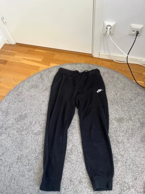 Svarta mjukisbyxor från Nike - Säljer ett par svarta mjukisbyxor från Nike med klassisk logga på vänster ben. Byxorna har dragsko i midjan och muddar vid bensluten. Perfekta för chill dagar eller träning. Tillverkade i mjukt material för maximal komfort.