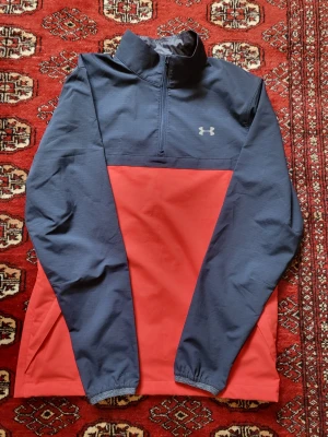 Blå och röd träningströja Under Armour - Snygg långärmad träningströja från Under Armour i blått och rött. Tröjan har halv dragkedja framtill, hög krage och elastiska muddar. Perfekt för löpning eller gym. Tillverkad i ett lätt och funktionellt material.