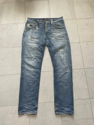 Blå slitna jeans med raka ben - Säljer ett par blå jeans från nudie i modellen STEADDY EDDIE med raka ben och tydliga slitningar samt hål på flera ställen. Jeansen är använda och lagade sen tidigare men är i väldigt fint skick! Snygga slitningar båda bak och fram samt en väldigt fin wash. Även en snygg slitning vid fickan. OBS, det finns ett litet hål vid gylfen men som går att laga enkelt (Kan lagas av mig ifall köparen så skulle vilja det innan köpet). Tveka inte att ställa frågor☺️