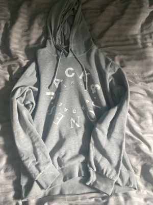 Grå hoodie från Jack & Jones - Snygg grå hoodie från Jack & Jones med stor vit logga tryckt framtill. Hoodien har huva med dragsko och ribbade muddar vid ärmar och nederkant. Mjuk insida för extra komfort, perfekt för chill dagar.