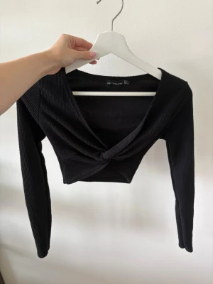 Svart croppad topp från PrettyLittleThing - Trendig svart croppad topp från PrettyLittleThing med lång ärm och twistad omlottdetalj framtill. Toppen är ribbad och har en snygg v-ringning. Använd enstaka gånger. 