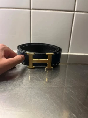 Mörkblå bälte med guldfärgat H-spänne - Snyggt svart bälte med ett stort guldfärgat H-spänne och vita kontrastsömmar. Bältet har en klassisk och stilren design som passar till många olika outfits. Perfekt accessoar för att lyfta din look.