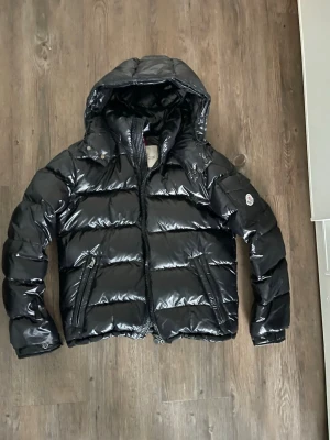 Svart glansig dunjacka från Moncler - Snygg svart dunjacka från Moncler med glansig finish och huva. Jackan har två dragkedjefickor framtill och Moncler-logga på ärmen. Perfekt för kalla vinterdagar och riktigt trendig look.