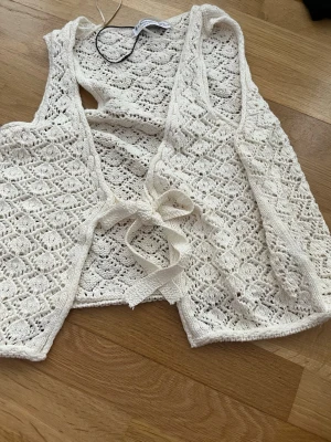 Vit stickad väst från Stradivarius - Säljer en söt vit stickad väst från Stradivarius i storlek S. Västen har ett fint virkat mönster och knytning framtill. Perfekt att slänga över en topp för en chill och trendig look.