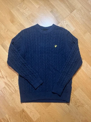 Lyle & Scott kabelstickad tröja - Säljer denna mörkblåa kabelstickad tröja med rund halsringning och ribbade muddar vid ärmslut och nederkant. Tröjan har ett gult broderat emblem på bröstet och är tillverkad i ett mjukt, tjockt material som passar perfekt för kyligare dagar. Klassisk och stilren design. Vid frågor kring varan eller fler bilder är det bara att skriva till mig!