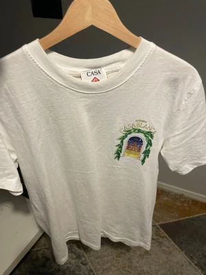 Vit t-shirt med tryck från Casa - Vit t-shirt från Casa med färgglatt tryck på bröstet där det står 'Avenida Casablanca' och ett motiv av en byggnad omgiven av gröna blad. Klassisk passform och rund halsringning. Perfekt för dig som gillar stilrena plagg med en twist.