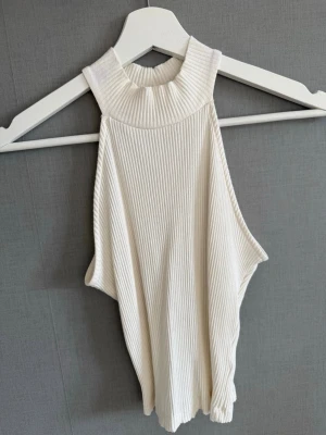 Vit ribbad ärmlös linnetopp - Säljer en vit ribbad linnetopp med hög hals och ärmlös design. Toppen har en croppad passform och är tillverkad i ett ,ä stretchigt material som sitter snyggt på kroppen. Använd en gång