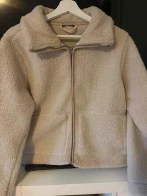 Beige teddyjacka med dragkedja - Mysig beige teddyjacka med stor krage och dragkedja framtill. Jackan har två stora fickor på framsidan och är gjord i fluffigt, mjukt material som håller dig varm. Perfekt för kyliga dagar och enkel att matcha med olika outfits.