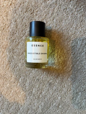 ESSNCE Irresistible Enigma parfym - En stilren parfymflaska från ESSNCE med doften Irresistible Enigma. Flaskan är rund i genomskinligt glas med svart lock och innehåller en guldfärgad vätska. Minimalistisk etikett framtill. Perfekt för dig som gillar unika dofter.