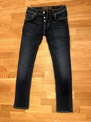 Jacob Cohën Jeans - Snygga mörkblå slim jeans med normal midja och fyra silverfärgade knappar i gylfen. Jeansen har klassiska fem fickor och kontrastsömmar. Materialet ser ut att vara stretchigt denim för en bekväm passform.