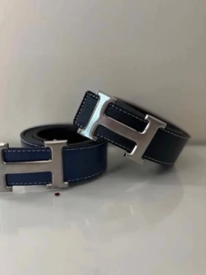 Mörkblått skärp med silverdetalj - Snyggt mörkblått bälte med stor silvrig H-formad spänne. Bältet har vita sömmar längs kanterna och är gjort i skinn. Perfekt accessoar för att lyfta din outfit.
