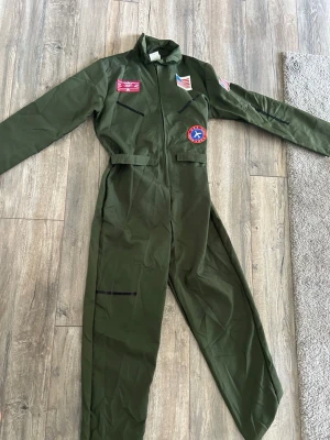Grön pilotdräkt - top gun - Cool grön pilotdräkt för maskerad med flera broderade patchar, bland annat amerikanska flaggor och en röd-blå pilotbadge. Dräkten har dragkedja framtill och ståkrage. Perfekt för dig som vill sticka ut på nästa temafest! Från filmserien Top Gun!
