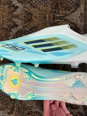 Adidas F50 fotbollsskor blå/vit - Säljer ett par Adidas F50 fotbollsskor i blått och vitt med gula detaljer. Skorna har en strömlinjeformad design utan snörning och en sula med iriserande finish och fasta dobbar. Perfekta för dig som vill ha lättviktsskor med grym känsla på planen.