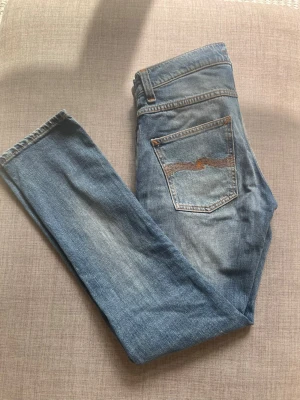 Blå jeans från Nudie Jeans Co - Säljer ett par klassiska blå jeans från Nudie Jeans Co i modellen W30 L32. Jeansen har en snygg tvätt, raka ben och den ikoniska vågade sömmen på bakfickan. Tillverkade i slitstark denim med orangea kontrastsömmar. Perfekta för en avslappnad och trendig look.