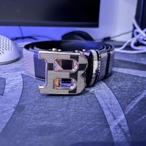  Burberry bälte - Snyggt bälte med breda svarta och gråa ränder och ett stort, silvrigt B-format spänne. Bältet har en stilren design och känns lyxigt med sitt blanka metallspänne. Perfekt accessoar för att lyfta din outfit med en cool detalj.