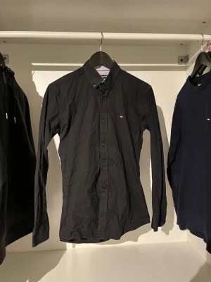 Svart skjorta från Tommy Hilfiger - Klassisk svart skjorta från Tommy Hilfiger med button-down krage och liten logga på bröstet. Skjortan har långa ärmar och knäppning framtill. Perfekt till jeans eller chinos för en clean och stilren look.