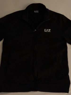 Svart ziptröja från EA7 Emporio Armani - Snygg svart ziptröja från EA7 Emporio Armani med vit logga på bröstet. Tröjan har hög krage, dragkedja framtill och långa ärmar. Perfekt för en sportig och stilren look. Sällan använd