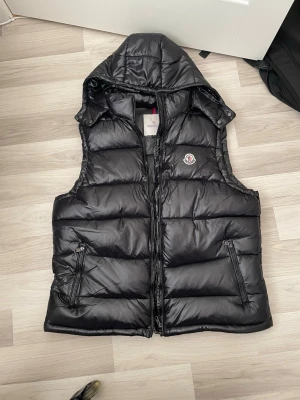  Moncler väst - Snygg svart dunväst från Moncler med glansig finish, avtagbar huva och två dragkedjeförsedda fickor. Klassisk Moncler-logga på bröstet och quiltad design som ger en modern look. Perfekt för lager-på-lager-stil under kyliga dagar.