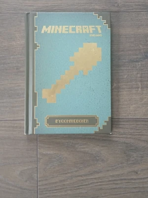Minecraft Bygghandboken bok - Minecraft Bygghandboken är en inbunden bok med blågrönt omslag och guldfärgade detaljer. Boken innehåller tips och steg-för-steg-instruktioner för att bygga i Minecraft, samt inspiration från communityn. Perfekt för dig som vill bli en mästerbyggare.