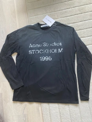 Svart långärmad topp Acne Studios S - Svart långärmad topp från Acne Studios med tryck 'Acne Studios STOCKHOLM 1996' i vitt framtill. Klassisk rund halsringning och rak passform. Perfekt för dig som gillar stilrena och trendiga plagg.