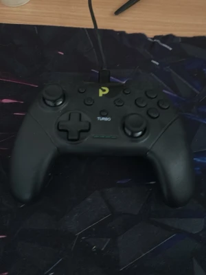 Svart trådbunden spelkontroll - En svart spelkontroll med ergonomisk form och tydliga knappar, inklusive styrkors, två styrspakar och TURBO-knapp. Kontrollen har en USB-kabel för anslutning och är tillverkad i plast. Passar perfekt för gaming på PC eller konsol.