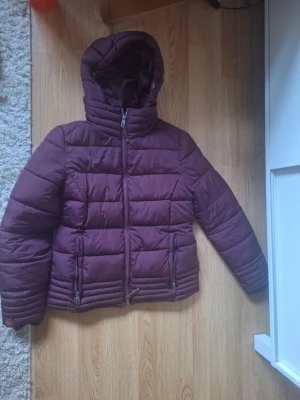 vinterjacka  - This is a women's or girl's puffer jacket in a plum or dark purple color. It features a hood, front zipper, and two zippered pockets. The jacket has a quilted design for warmth. The brand 'Marikoo' is shown on a brown patch on the sleeve, with 'EST 2016' also visible.