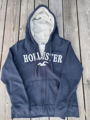 Marinblå hoodie från Hollister - Mysig marinblå hoodie från Hollister med dragkedja, stor vit logga på bröstet och fluffigt teddyfoder i huvan. Hoodien har fickor framtill och justerbara snören vid huvan. Perfekt för dig som gillar en avslappnad och varm stil.