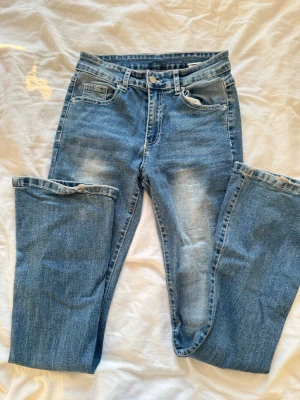 Blå bootcut jeans med broderade fickor - Säljer ett par blå bootcut jeans med snygg slitning framtill och broderade bakfickor med knappar. Jeansen har klassisk femficksmodell och normalhög midja. Perfekta för dig som gillar en avslappnad men trendig look.